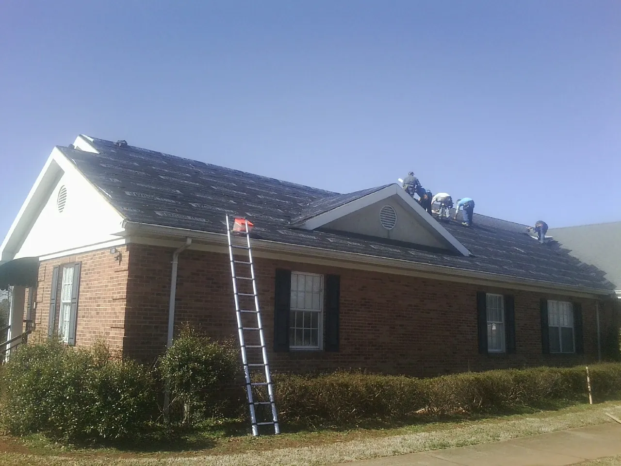 Crew tearing shingles off a brick ranch roof with a ladder set — Metal Roof Repair work by our crew in Rural Hall