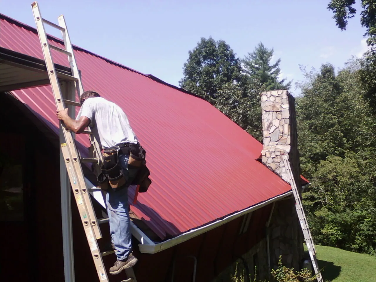 Roofer on a ladder beside a red metal roof and stone chimney — Soffit & Fascia Repair by Roofing Brothers in Rural Hall